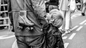 A young boy looks up at an adult, possibly a parent, who has a roller skate hanging over their shoulder. The scene takes place on a street with blurred people in the background, suggesting a busy or crowded environment.