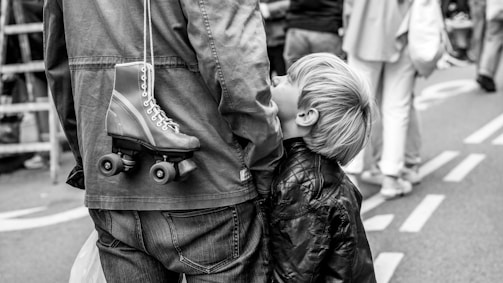 A young boy looks up at an adult, possibly a parent, who has a roller skate hanging over their shoulder. The scene takes place on a street with blurred people in the background, suggesting a busy or crowded environment.