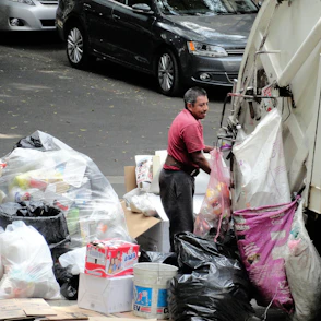 A person is seen working with a garbage truck, handling bags filled with various types of waste. The setting appears to be on a street with parked cars and a large pile of trash bags, boxes, and other debris. The individual is wearing a red shirt and is standing next to the truck, possibly involved in waste management or collection.