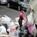 A person is seen working with a garbage truck, handling bags filled with various types of waste. The setting appears to be on a street with parked cars and a large pile of trash bags, boxes, and other debris. The individual is wearing a red shirt and is standing next to the truck, possibly involved in waste management or collection.