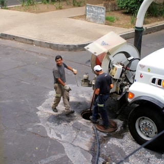 Two workers are engaged in maintenance work on a street, using a specialized vehicle equipped with a hose and large equipment. One worker is handling tools near an open manhole, while the other operates machinery on the vehicle.