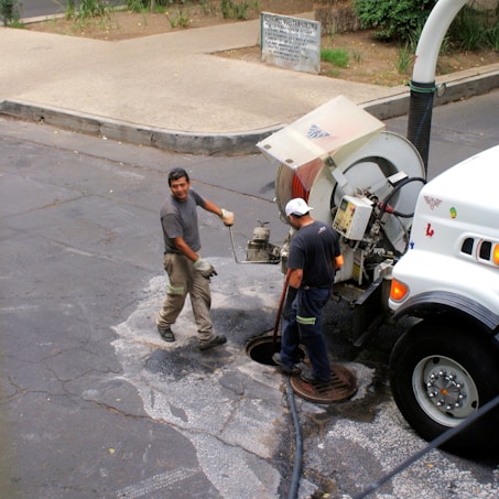 Two workers are engaged in maintenance work on a street, using a specialized vehicle equipped with a hose and large equipment. One worker is handling tools near an open manhole, while the other operates machinery on the vehicle.
