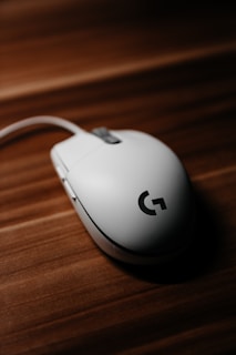 A white computer mouse with a prominent logo is placed on a wooden surface. The lighting highlights the smooth curves and design of the mouse, creating a contrast with the textured background.
