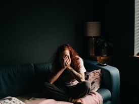 A person with long, curly hair is seated on a couch with their hands covering their face, creating a sense of introspection or distress. The room is dimly lit, with a lamp on a wooden side table next to a plant. A remote control rests on the couch. The overall atmosphere is one of solitude.