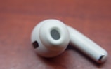 A single white wireless earbud is placed on a smooth, reddish-brown surface. The earbud's speaker mesh and charging connectors are visible.