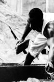 A person is engaged in manual labor, holding a brick while wearing a sleeveless shirt and shorts. The scene appears to be part of a construction site, with blurred structures in the background. The image is in black and white, emphasizing the contrast and details of the person's muscles and the texture of the brick.