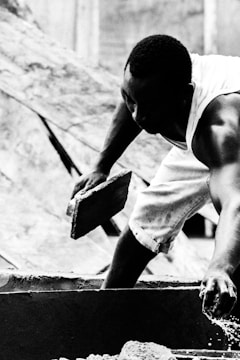A person is engaged in manual labor, holding a brick while wearing a sleeveless shirt and shorts. The scene appears to be part of a construction site, with blurred structures in the background. The image is in black and white, emphasizing the contrast and details of the person's muscles and the texture of the brick.