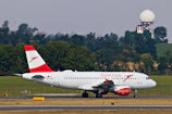 A commercial airplane with the Austrian Airlines logo is taxiing on a runway with a backdrop of trees and a radar tower in the background. The aircraft is white with red accents, visible registration number OE-LDG, and features distinct branding on the fuselage and engine.
