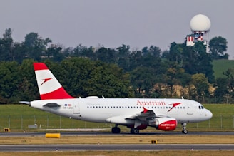 A commercial airplane with the Austrian Airlines logo is taxiing on a runway with a backdrop of trees and a radar tower in the background. The aircraft is white with red accents, visible registration number OE-LDG, and features distinct branding on the fuselage and engine.