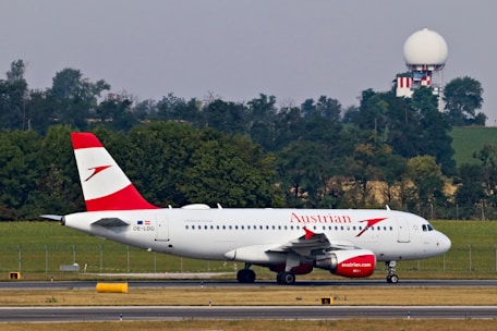 A commercial airplane with the Austrian Airlines logo is taxiing on a runway with a backdrop of trees and a radar tower in the background. The aircraft is white with red accents, visible registration number OE-LDG, and features distinct branding on the fuselage and engine.