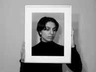 A person wearing a black top holds an empty photo frame around their face. The scene is captured in black and white, adding a classic and introspective feel. The person looks slightly to the side, blending with the frame in a way that creates a portrait within a portrait effect.