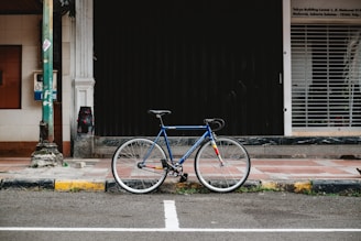 A blue bicycle with a fixed-gear stands against a black shuttered storefront on a sidewalk. The setting is urban, with tiled pavement and a worn, yellow-painted curb. A utility pole is visible on the left with a small box attached to it. Behind the pole, there is a white column next to a brown-tiled section of the building.
