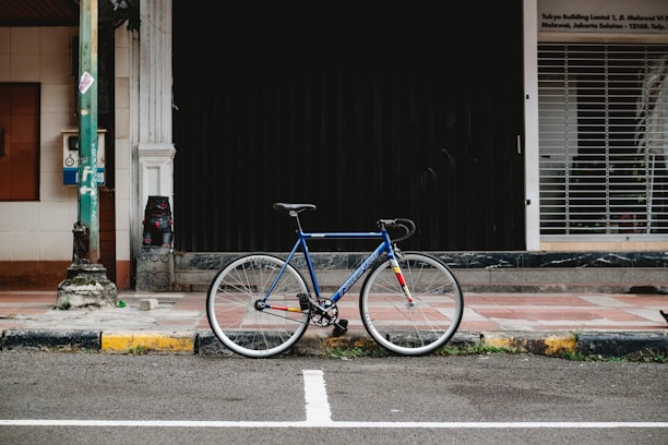 A blue bicycle with a fixed-gear stands against a black shuttered storefront on a sidewalk. The setting is urban, with tiled pavement and a worn, yellow-painted curb. A utility pole is visible on the left with a small box attached to it. Behind the pole, there is a white column next to a brown-tiled section of the building.