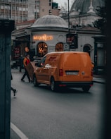 A bustling city street scene featuring an orange DHL delivery van parked in front of a building with a distinctive domed architecture. Several people, including a delivery person handling a package, are visible near the van. The illuminated sign for The House Cafe can be seen on the building.