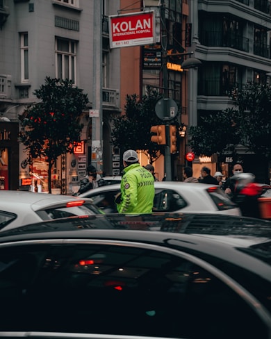 A busy urban street scene featuring a traffic police officer in a reflective vest directing vehicles. Tall buildings and storefronts line the street, with pedestrians walking by. Cars are stopped, and the street is bustling with activity. A sign for Kolan International Hospital is visible above.