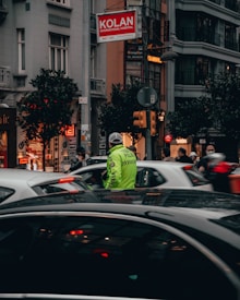 A busy urban street scene featuring a traffic police officer in a reflective vest directing vehicles. Tall buildings and storefronts line the street, with pedestrians walking by. Cars are stopped, and the street is bustling with activity. A sign for Kolan International Hospital is visible above.