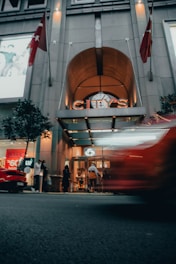 An entrance to a modern building, City’s, with illuminated signage and glass doors. People are gathered near the entrance, with a sale sign in the background. A blurred red car is passing by on the street in the foreground, and there are flags on both sides of the building's facade.