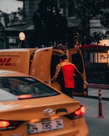 A person wearing a bright red and yellow uniform stands next to an open yellow delivery van, possibly loading or unloading packages. In the foreground, a yellow taxi is partially visible, adding to the urban street setting. Surrounding buildings and trees are visible, contributing to a busy city atmosphere.