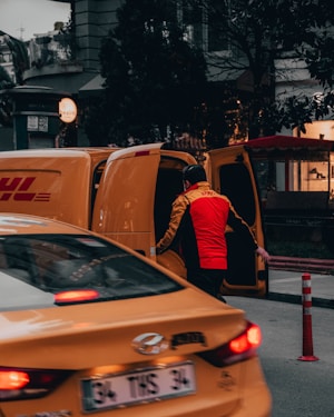 A person wearing a bright red and yellow uniform stands next to an open yellow delivery van, possibly loading or unloading packages. In the foreground, a yellow taxi is partially visible, adding to the urban street setting. Surrounding buildings and trees are visible, contributing to a busy city atmosphere.