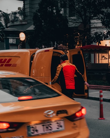 A person wearing a bright red and yellow uniform stands next to an open yellow delivery van, possibly loading or unloading packages. In the foreground, a yellow taxi is partially visible, adding to the urban street setting. Surrounding buildings and trees are visible, contributing to a busy city atmosphere.