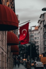 A street scene in a city with a Turkish flag prominently displayed on a pole. The buildings are adorned with red canopy awnings. Cars and a few pedestrians are visible on the road and sidewalks. The sky is cloudy, and the overall lighting suggests an overcast day.