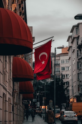A street scene in a city with a Turkish flag prominently displayed on a pole. The buildings are adorned with red canopy awnings. Cars and a few pedestrians are visible on the road and sidewalks. The sky is cloudy, and the overall lighting suggests an overcast day.