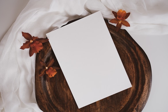 A blank white rectangular card is placed on a wooden tray. Surrounding the tray are four brownish-red flowers resting on a white fabric. The setup creates a natural and earthy aesthetic.
