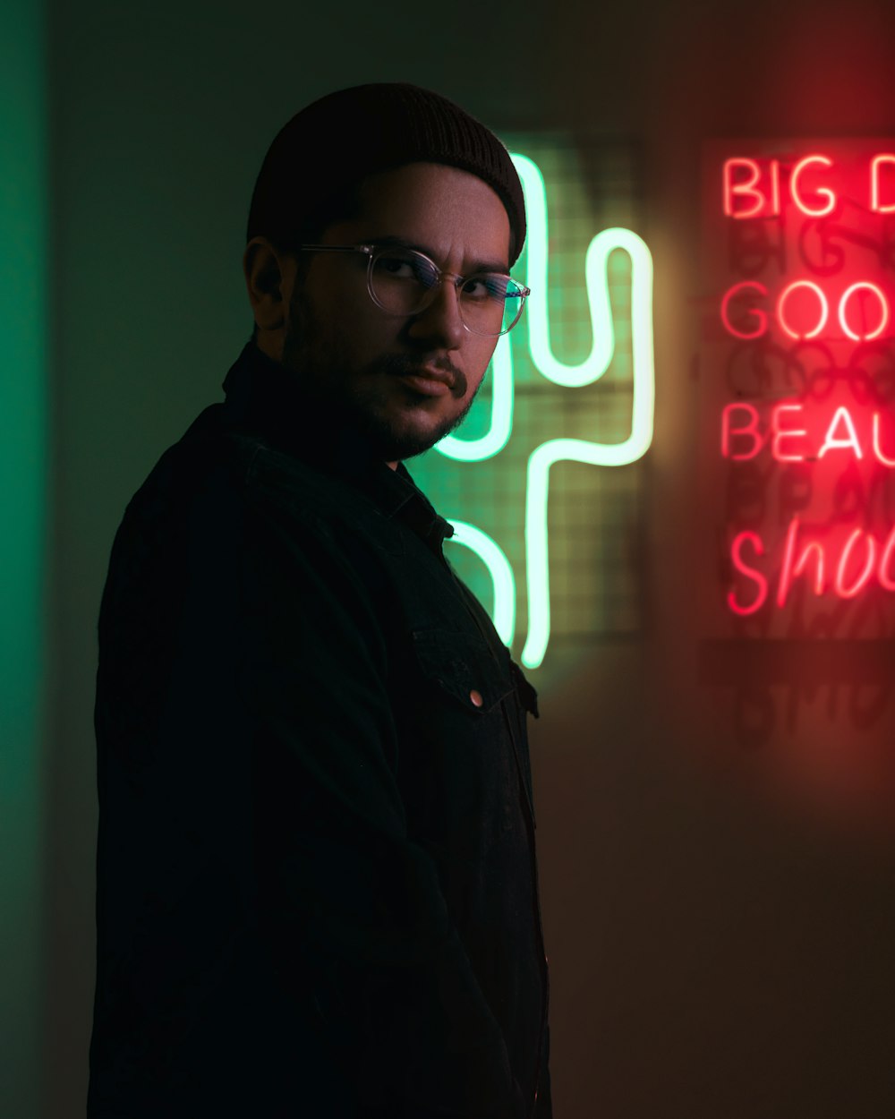 man in black jacket wearing black knit cap standing near red neon signage