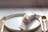 A neatly arranged table setting featuring a white ceramic plate, a beige cloth napkin rolled and tied with twine, adorned with small pink flowers. The setup is placed on a woven placemat with golden cutlery on either side, resting on a light-colored textured surface.