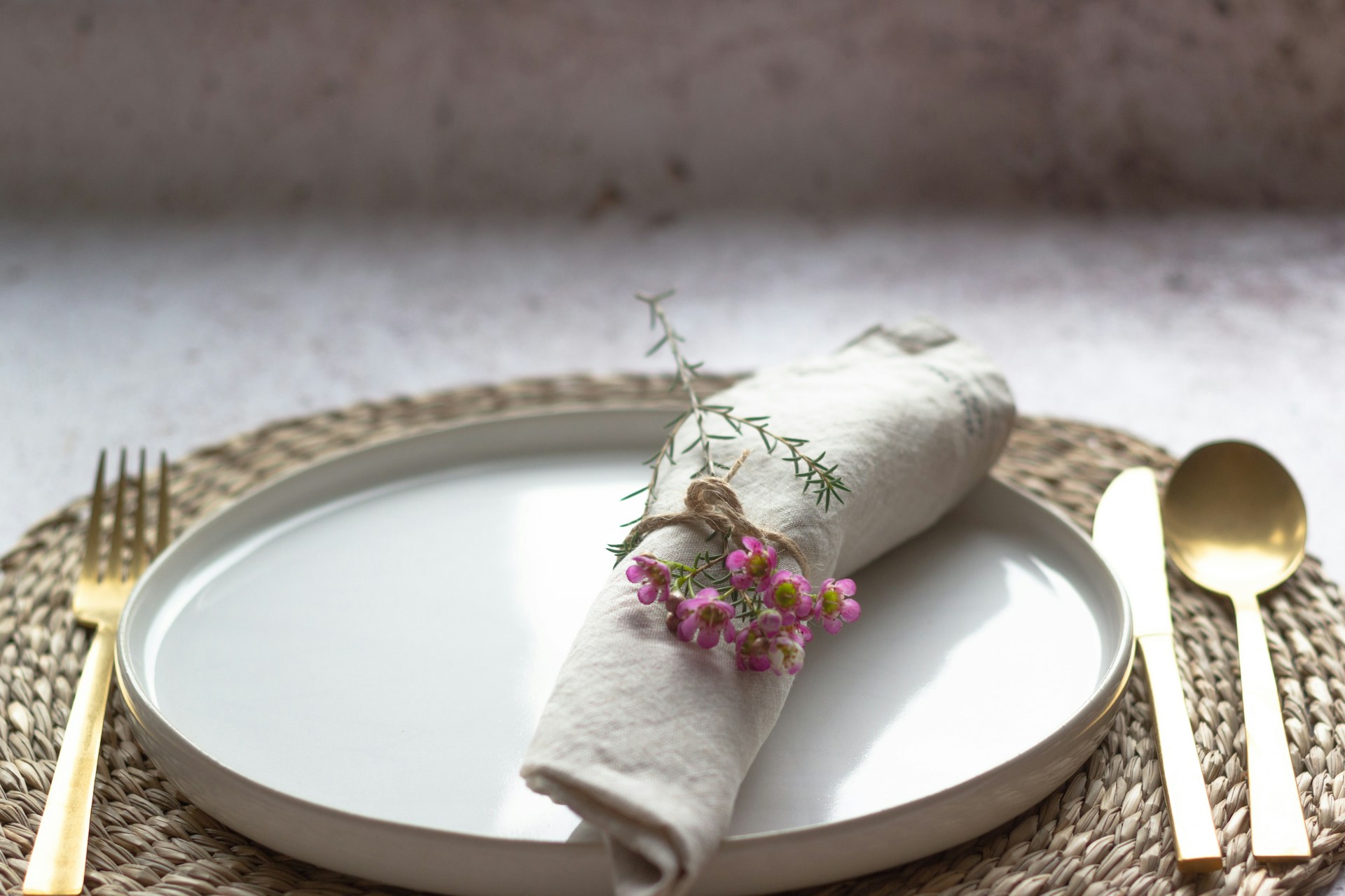 A neatly arranged table setting featuring a white ceramic plate, a beige cloth napkin rolled and tied with twine, adorned with small pink flowers. The setup is placed on a woven placemat with golden cutlery on either side, resting on a light-colored textured surface.