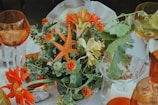 A detailed and colorful floral arrangement featuring vibrant orange and yellow flowers, including sunflowers and marigolds, mixed with green foliage. A starfish is creatively incorporated into the display. The arrangement is set on a table with an elegant white tablecloth, surrounded by amber-hued glassware. Leafy greens add texture, and a candle in a glass holder adds a subtle touch of warmth.