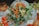 A detailed and colorful floral arrangement featuring vibrant orange and yellow flowers, including sunflowers and marigolds, mixed with green foliage. A starfish is creatively incorporated into the display. The arrangement is set on a table with an elegant white tablecloth, surrounded by amber-hued glassware. Leafy greens add texture, and a candle in a glass holder adds a subtle touch of warmth.
