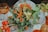A detailed and colorful floral arrangement featuring vibrant orange and yellow flowers, including sunflowers and marigolds, mixed with green foliage. A starfish is creatively incorporated into the display. The arrangement is set on a table with an elegant white tablecloth, surrounded by amber-hued glassware. Leafy greens add texture, and a candle in a glass holder adds a subtle touch of warmth.