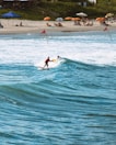 Two surfers ride a wave near a sandy beach lined with colorful umbrellas and sunbathers. The ocean is a vibrant blue, and the beach is bustling with people relaxing under the shade. Red flags are placed in the sand and water, indicating possible surf conditions. The scene is lively, capturing a typical day at a popular beach destination.