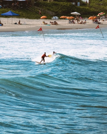 Two surfers ride a wave near a sandy beach lined with colorful umbrellas and sunbathers. The ocean is a vibrant blue, and the beach is bustling with people relaxing under the shade. Red flags are placed in the sand and water, indicating possible surf conditions. The scene is lively, capturing a typical day at a popular beach destination.