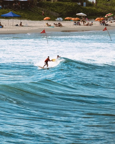 Two surfers ride a wave near a sandy beach lined with colorful umbrellas and sunbathers. The ocean is a vibrant blue, and the beach is bustling with people relaxing under the shade. Red flags are placed in the sand and water, indicating possible surf conditions. The scene is lively, capturing a typical day at a popular beach destination.