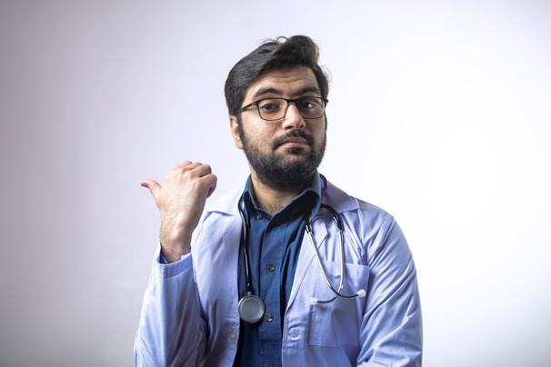 A man with a beard and glasses is wearing a white coat and a stethoscope around his neck. He is gesturing with his thumb pointing to the side and has a confident and thoughtful expression.