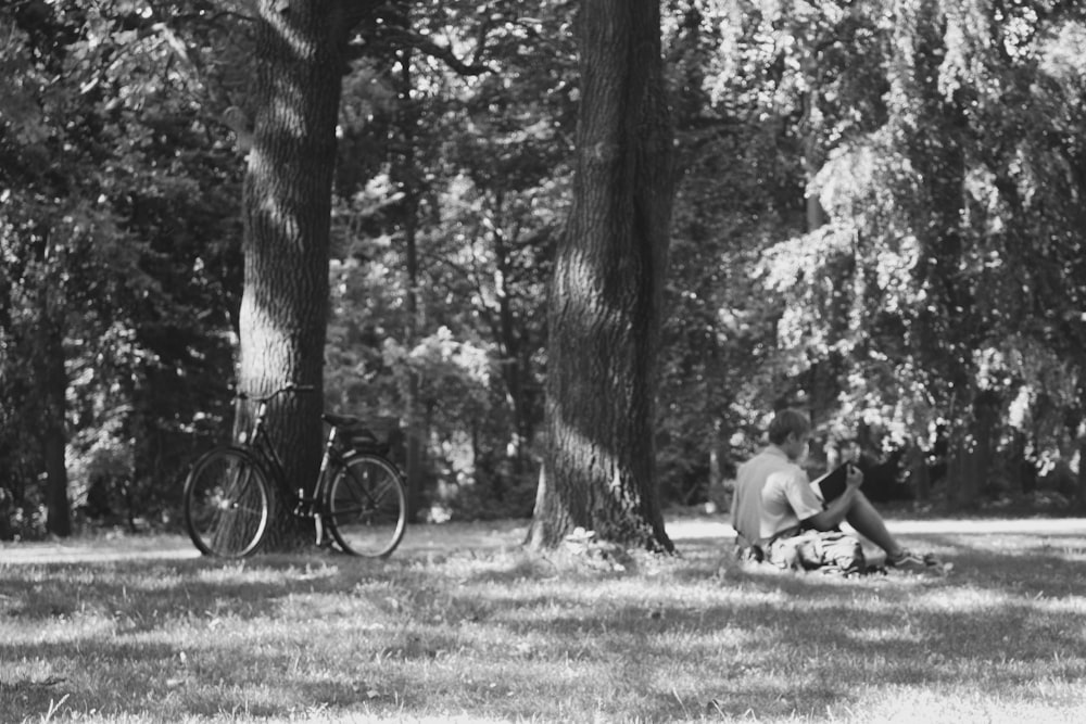grayscale photo of man sitting on grass field near tree
