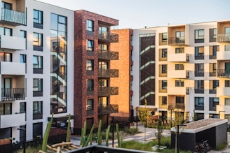 A modern apartment complex with multiple buildings. The buildings have contemporary architectural designs and are painted in various colors, including white, beige, and red brick. Several balconies and large windows are visible on each building. The courtyard area contains green plants, small trees, and a pathway. There's also a structure at the bottom right corner that might be a storage or utility shed.