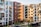 A modern apartment complex with multiple buildings. The buildings have contemporary architectural designs and are painted in various colors, including white, beige, and red brick. Several balconies and large windows are visible on each building. The courtyard area contains green plants, small trees, and a pathway. There's also a structure at the bottom right corner that might be a storage or utility shed.
