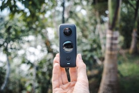 A hand holds a 360-degree camera vertically in an outdoor setting, surrounded by blurred trees and natural foliage in the background. The device features two lenses, one on top and another in the middle, capturing a full panoramic view.