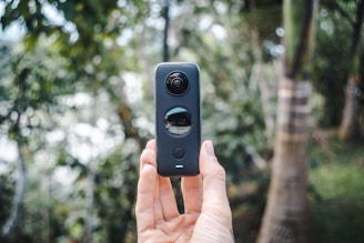 A hand holds a 360-degree camera vertically in an outdoor setting, surrounded by blurred trees and natural foliage in the background. The device features two lenses, one on top and another in the middle, capturing a full panoramic view.