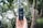 A hand holds a 360-degree camera vertically in an outdoor setting, surrounded by blurred trees and natural foliage in the background. The device features two lenses, one on top and another in the middle, capturing a full panoramic view.