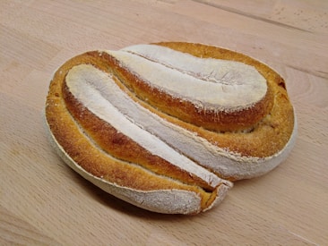 A freshly baked loaf of bread with a golden brown crust, displayed on a light wooden surface. The loaf has a rustic appearance with a floured top and distinct slashes on the crust.