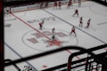 A group of ice hockey players is positioned on a rink with a large logo at the center. Most players are wearing red uniforms and helmets, and are preparing for a face-off or are practicing. The scene is viewed from a higher angle, suggesting it's taken from the stands. Surrounding advertisements line the rink boards.