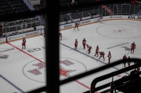 A group of ice hockey players wearing red uniforms is seen on the rink spread out in formation, likely part of a team lineup or practice session. The arena appears to be relatively empty, with a few spectators visible in the stands. Advertisements in Russian line the surrounding boards of the rink.