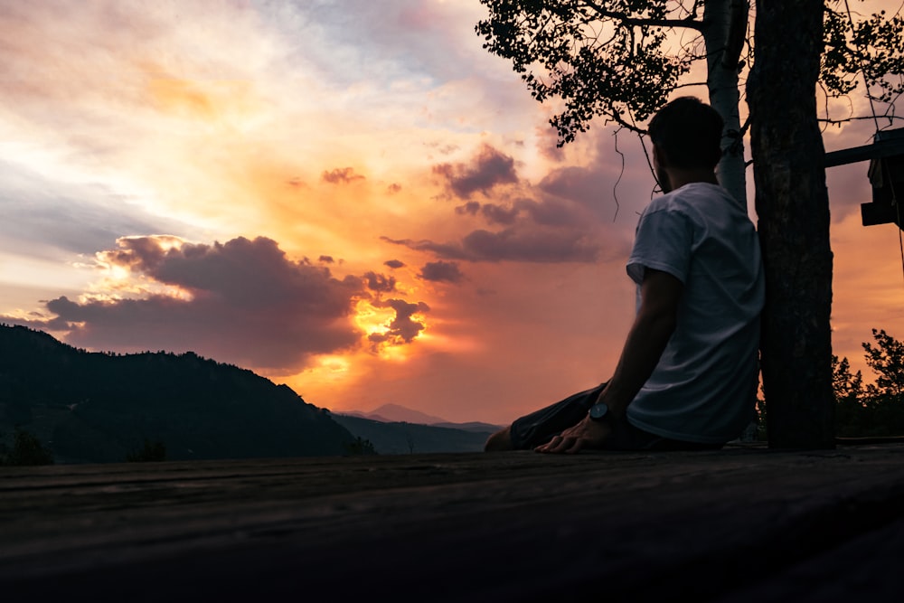 man in white t-shirt sitting on brown tree branch during sunset