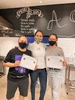 Three people stand close together, each wearing a mask. Two of them hold certificates, and the person in the middle wears a chef's jacket. The background features a blackboard with handwritten text and a kitchen setting.