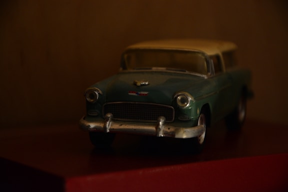 A vintage toy car model, mainly in green and silver with a classic design, placed on a dark surface against a blurred background. It appears to be a miniature of a classic sedan, capturing the details of the headlights and front grille.