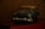 A vintage toy car model, mainly in green and silver with a classic design, placed on a dark surface against a blurred background. It appears to be a miniature of a classic sedan, capturing the details of the headlights and front grille.